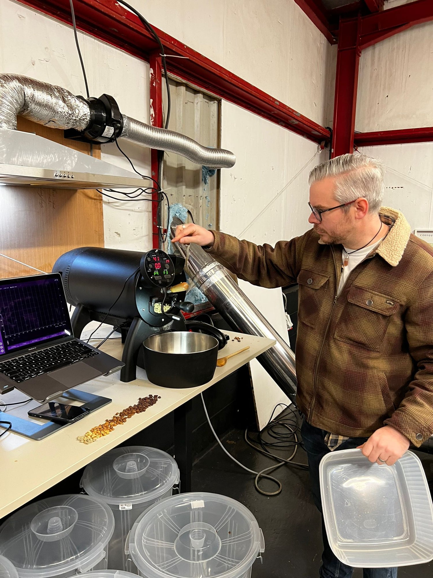 Chris in a brown jacket stands in a Danelaw Coffee Roastery, holding a clear plastic box while operating a Allio Bullet Coffee Roaster on a table with a laptop and containers of coffee nearby.