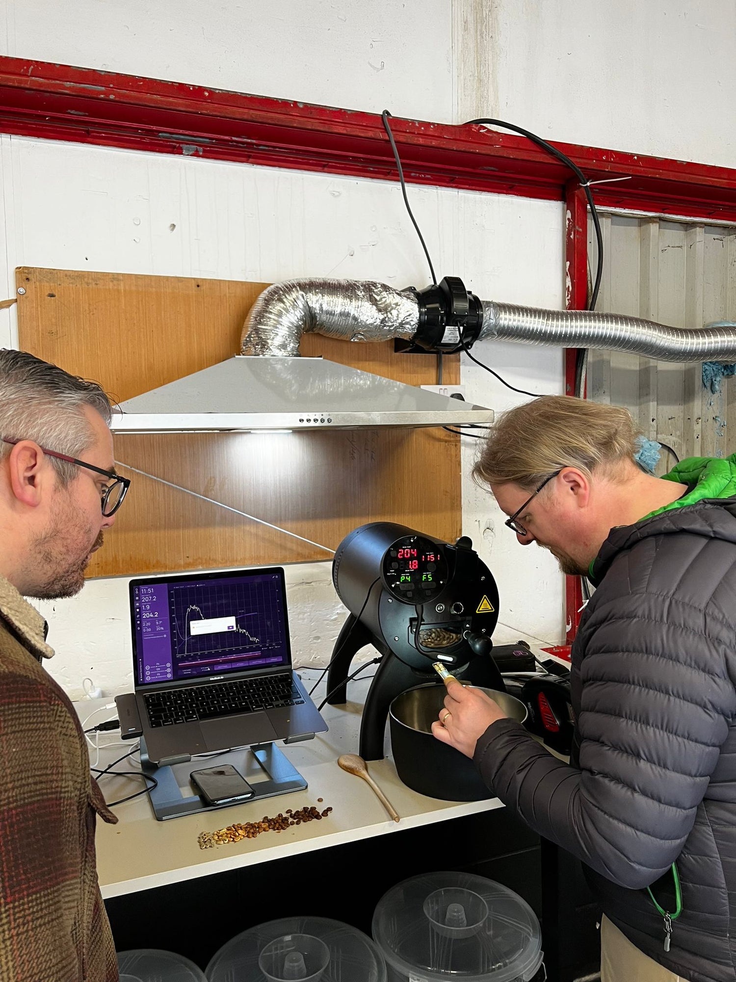 Chris and Dave at Danelaw Coffee in a roastery, one operating a coffee roasting machine Allio Bullet, while monitoring a laptop screen showing temperature and graph data.