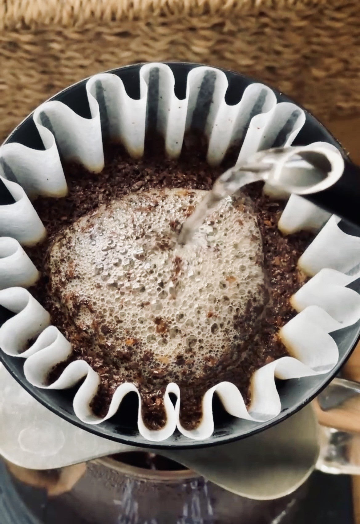 A close-up of a pour-over coffee setup with dark coffee grounds in a white paper filter inside a black cup, with hot water being poured from a black kettle.
