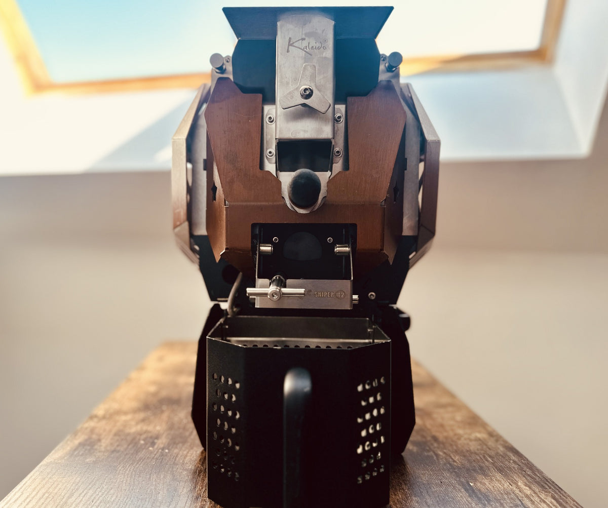 A Kaleido Sniper M2 Pro Coffee Roaster coffee roaster photographed from the front, sitting on a wooden table in the roastery with a skylight in the background.