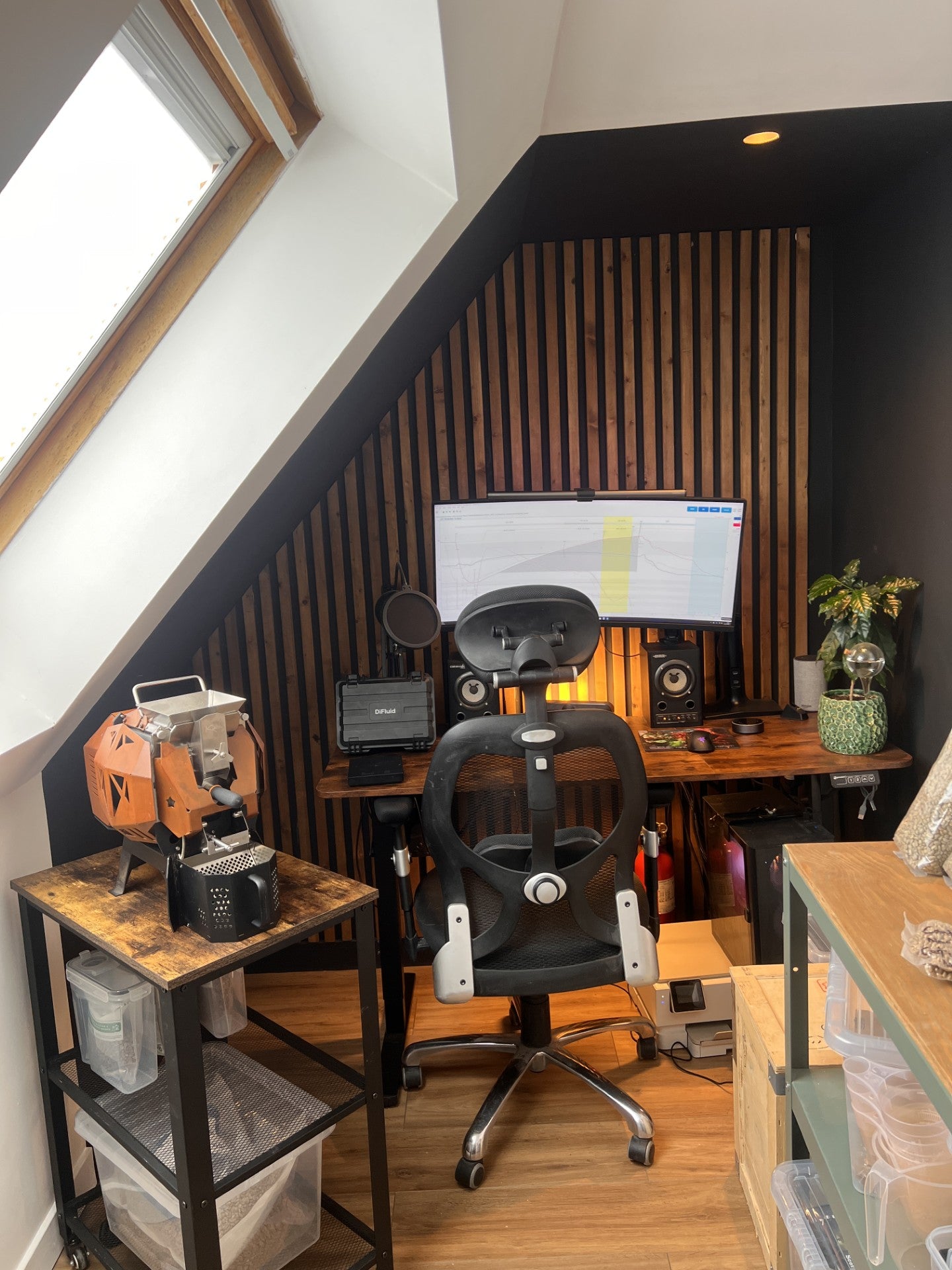 The roastery setup in a slanted upstairs room, featuring a black ergonomic office chair in front of a desk with a curved monitor, speakers, and a computer setup. The background wall has vertical wood slats, and the room has a wood floor and a large window on the left, coffee plant on the desk and coffee roaster under the window, with a worktop and bags of green beans on it.